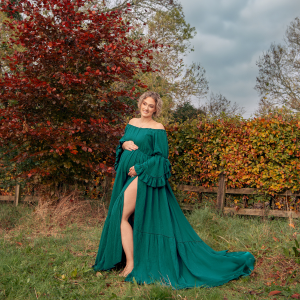 Outdoors maternity photoshoot. Woman holds her bump, in a green long flowing dress with a slit up one leg